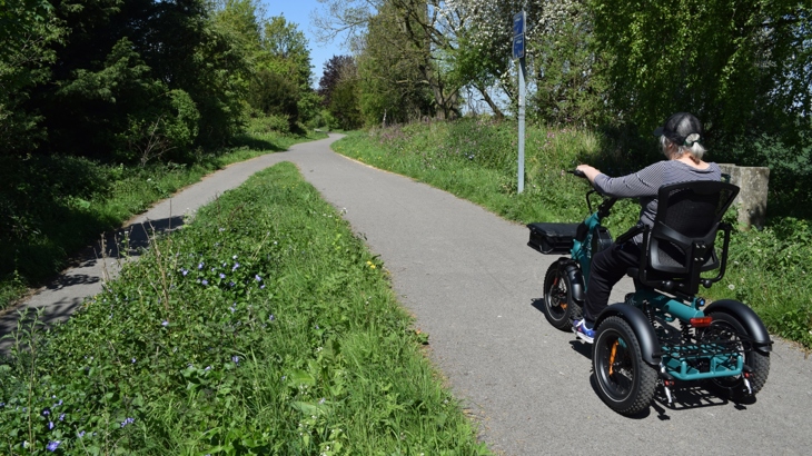 A person traveling along a national cycle network path on an electric trike. Around the path is lost of greenery.