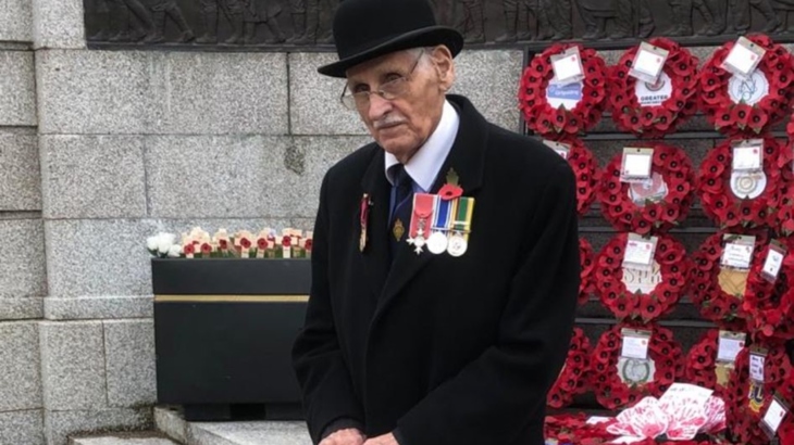 A man with glasses and a moustache wearing a coat adorned with miliary medals, a bowler hat and carrying an umbrella stands in front of poppy wreaths at a war memorial.stands 