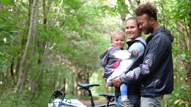 A father and a mother embracing in a woodland setting while holding their toddler the family are stood in front of their bikes and smiling