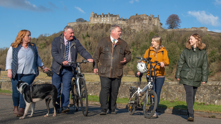 A group walking with Stirling Castle in the background. From left to right: Clackmannshire Council Leader Cllr Ellen Forson with dog Loki, Stirling Council Leader Cllr Scott Farmer, Stirling Council Depute Leader Cllr Chris Kane, Walk Wheel Cycle Trust Portfolio Director for Scotland Karen McGregor, Environment Advisor at the University of Stirling Amy Gove-Kaney.