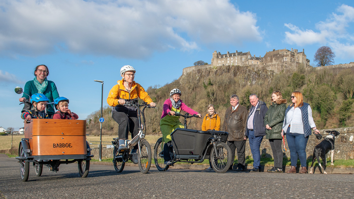 Image of a group of cyclists with Stirling Castle as the backdrop. On the left there is a cargo bike with two small children in it, in the middle a man rides a small push bike and to his right is another cargo bike.