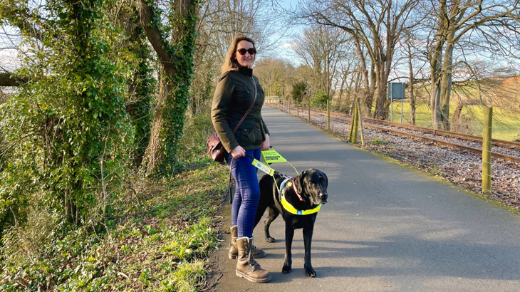 A woman called Sarah poses for a photograph with her black Labrador guide dog called Webbly. Webbly's wearing a guiding harness which Sarah's holding. The pair are on the Bristol Bath Railway Path, National Route 4 on a sunny spring day.