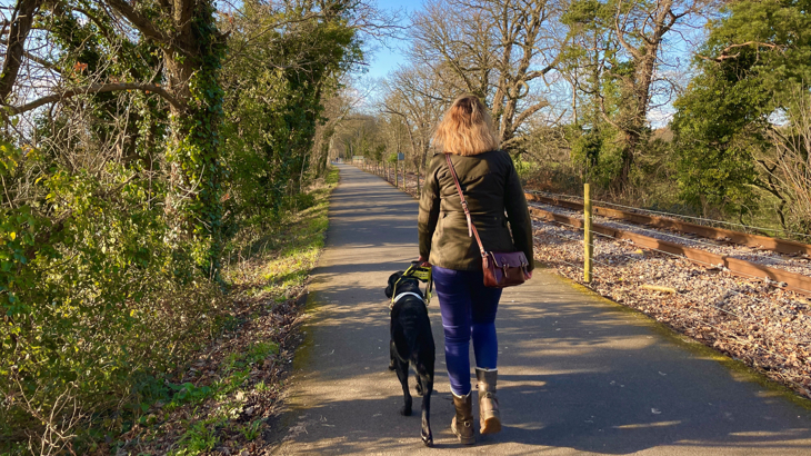 A woman called Sarah walks away from the camera with her black Labrador guide dog called Webbly. Webbly's wearing a guiding harness which Sarah's holding. The pair are on the Bristol Bath Railway Path, National Route 4 on a sunny spring day.