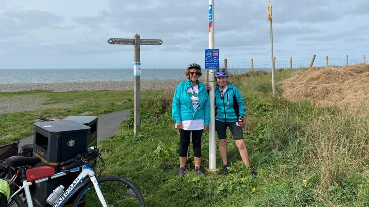 Two women stood next to each other next to a National Cycle Network sign on a cloudy day by the coast in the UK. The photograph of the two friends was taken during their 80-mile cycle on the Bay Cycle Way in Lancaster.