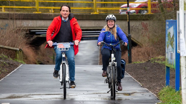A man and a woman looking happy cycling front on along a new path with fresh-looking tarmac. In the background there's a small bridge with a car passing over. 