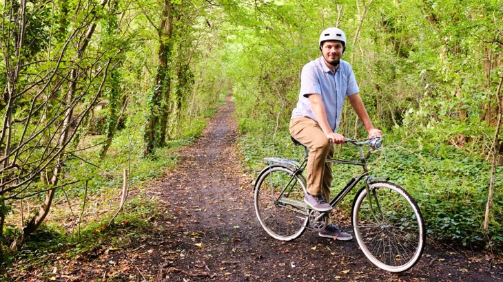 An adult male stops on their bike on the Lias Line Greenway in Warwickshire. Behind them, the path is a leafy corridor of trees and foliage on a bright day.