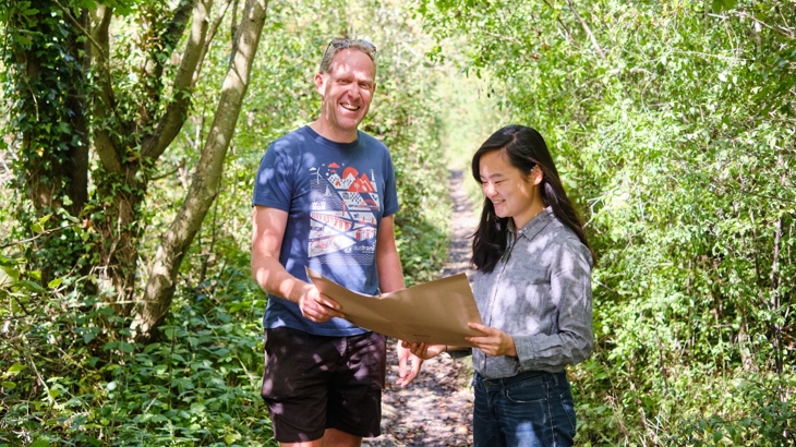 Two Walk Wheel Cycle Trust colleagues stand on the Lias Line Greenway in Warwickshire, looking over plans for the path's future development. The scene behind them is a narrow, leafy corridor of trees and foliage on a sunny day.