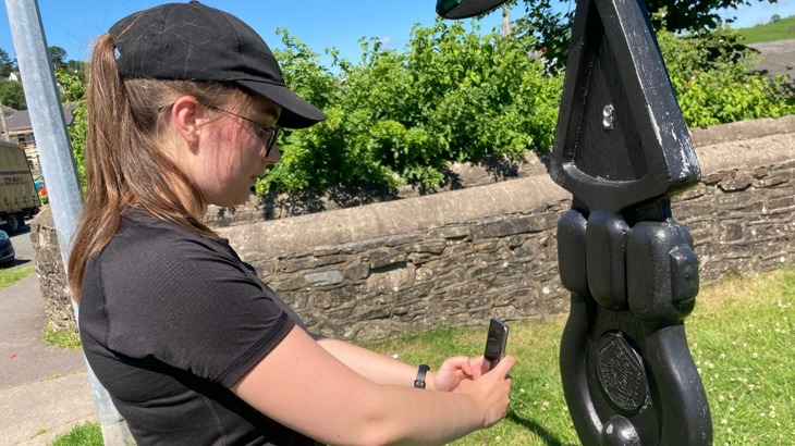 A young volunteer with a black baseball cap on taking a photo of a Millennium Milepost using her smartphone on a sunny day
