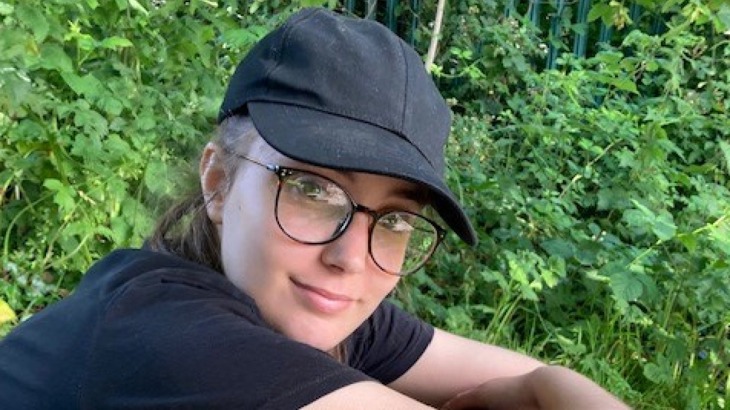 A closeup of a young person with glasses and a black baseball hat on smiling at the camera