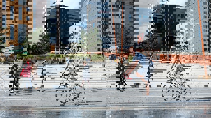 city plaza with fountains and people walking and cycling 