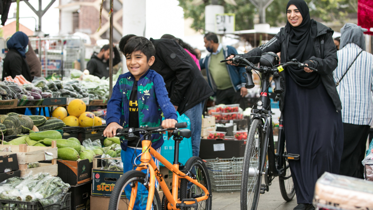 A child and and woman smiling while walking their bicycles through a busy fruit and vegetable market 