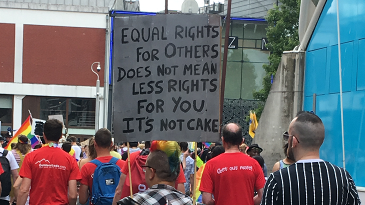 A large, hand painted, overhead banner at a busy Pride march reads 'EQUAL RIGHTS FOR OTHERS DOES NOT MEAN LESS RIGHTS FOR YOU. IT'S NOT CAKE'. It is written in black paint on card which is held up by garden canes. In the crowd are numerous rainbow flags and in the foreground, a person with rainbow dyed short hair.
