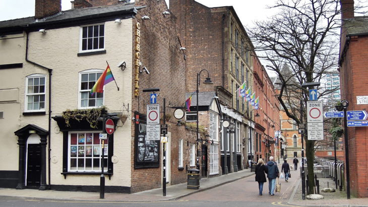 Canal Street in Manchester on a winter's day. The trees which line the street are bare, pedestrian zone signs are either side of the entrance to the street, a couple walk arm in arm and other people walk in the distance. In the foreground, an old brick built pub called Churchills flies a large pride flag. More flag can be seen further down the street.