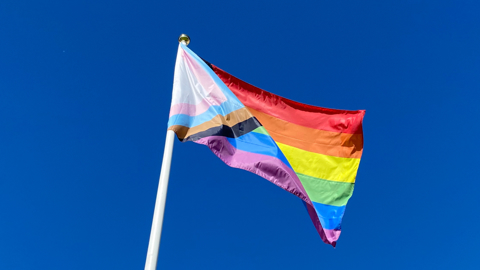 A Progress Pride flag on a flag pole flies in the wind against a bright blue sky.