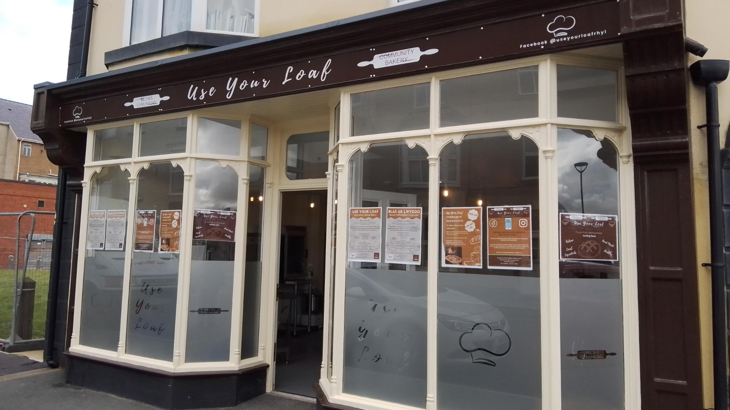 The exterior of a bakery called Use Your Loaf. The shop front has bay windows either side of the front door. The glazing is frosted to the lower half with the bakery name and the image of a chef's hat cut from the frosting. In the upper parts of the windows are a range of posters in brand tan and brown colours.