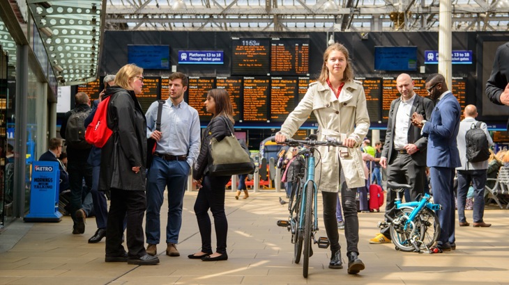 A woman commuter walks through a busy train station with her bike as live schedule boards are lit up behind her