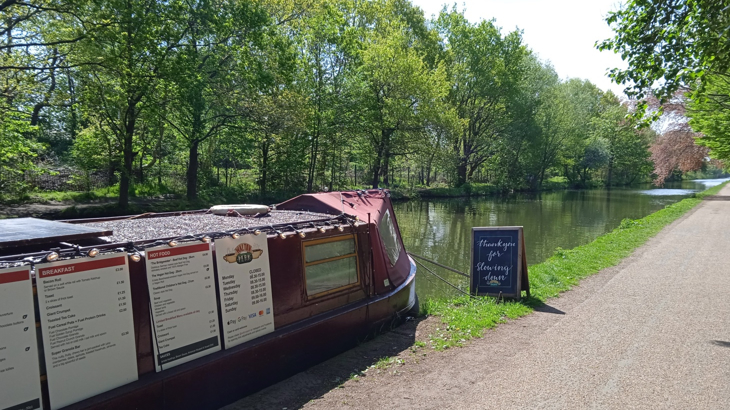A canal boat with a cafe inside floating on the water next to a path which is dotted with green trees just outside of Manchester, England