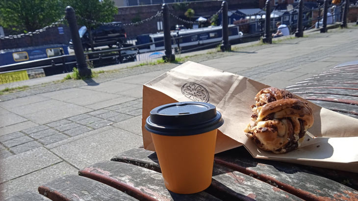 A takeaway cup and a baked bun perched on an outdoor bench in the sunshine with a canal and canal boats in the background 