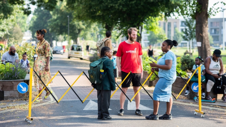 A group of people stand in front of a lattice gate across a road, which forms a temporary closure for cars during school drop-off and pick-up times. The sun is shining, the people are smiling and the mood is happy. 