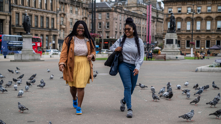 Two young women walking through a square in George Square in Glasgow with pigeons dotted around on the ground
