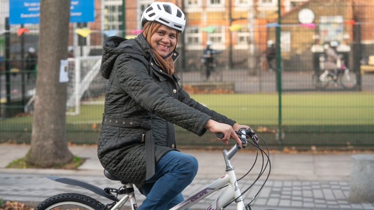 A woman rides a bicycle wearing a headscarf and cycle helmet. She is smiling and the sun is shining. In the background is a school.
