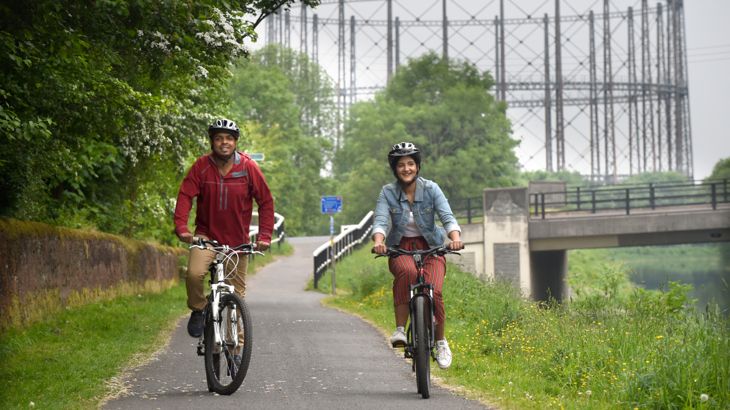 A man and a woman smiling cycling along a path with greenery either side on National Cycle Network Route 754 at Bowling Basin and Maryhill Locks, Glasgow
