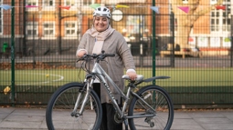 A woman wearing a headscarf under a cycle helmet stands in a school playground holding a bicycle. She is smiling, the autumn sun is shining, the mood is happy. 