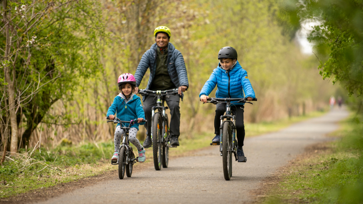 Adult male with two primary age children cycle on a traffic-free path. The straight path is tree lined and the day looks cool and dry. The group are wearing jackets and helmets.