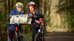 Two women are stopped on their mountain bikes on a traffic-free path in a woodland. They are looking at a map and smiling. They are wearing helmets and cycling clothing.