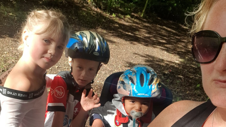 A woman stood next to her three young children in a woodland area, her youngest child is sat on a bike seat with a bike helmet on