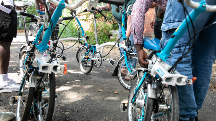 View from the ground of lots of legs and Brompton wheels on a sunny day. Everyone is stood with their bikes, some are folded.
