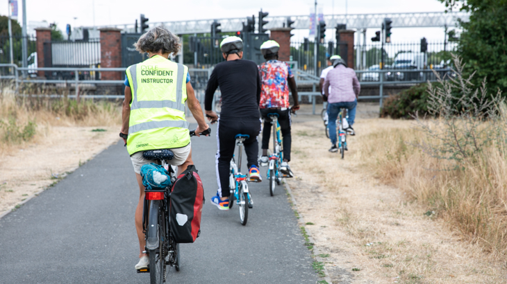 The view behind five people cycling on a traffic-free path. Four are on Brompton bikes, the other is on a standard bike with pannier. This person is wearing a hi-vis vest which says 'Cycle Confident Instructor'. Dry summer grass is either side of the sealed surface path and the group are approaching a road junction with traffic lights and a built up urban area beyond.