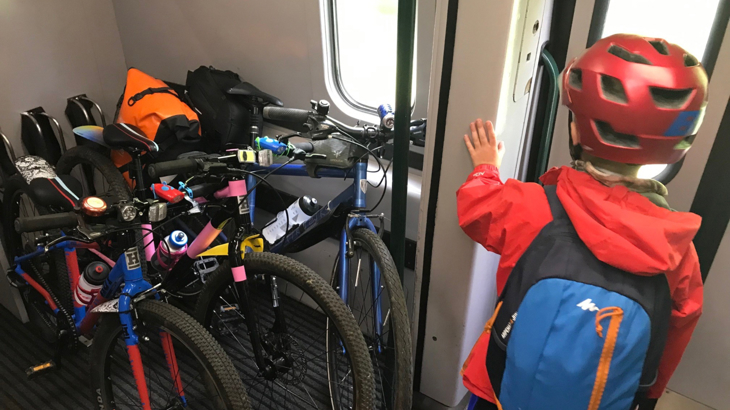 View of a small child from behind. They are wearing a red helmet, red anorak and blue backpack. They are stood in a train carriage, waiting at the closed door. Beside them is a cycle rack with three bikes loaded up with bags.