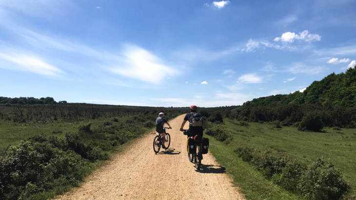 Adult and child cycling on an unsealed gravel path, a section of National Cycle Network towards Sway. The scenery is rural with rough, open green land to either side of the path and trees are in the distance. The sky is bright blue with occasional clouds. It looks like a hot day.