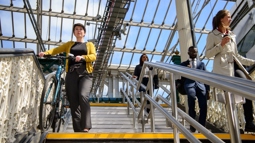Four commuters in formal office wear walk down a staircase in a glass roofed railway station. One is wheeling a bike on a custom bike ramp which runs parallel to the stairs. The sky beyond the glass ceiling is bright and blue.