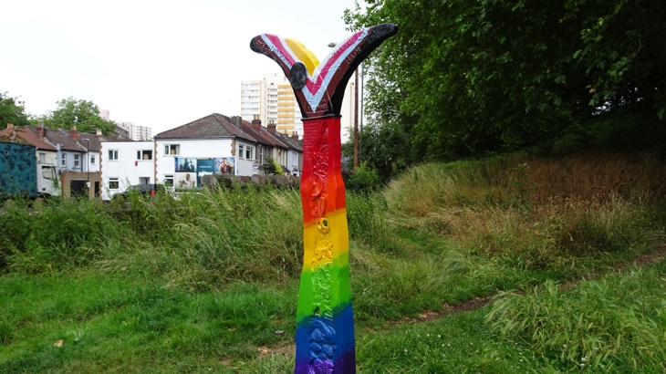 A milepost on the National Cycle Network in Bristol, painted in pride rainbow colours. Green park space and the city can be see in the background.