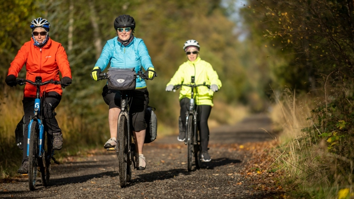 Three adult females on bikes, wearing helmets, coats and gloves, cycling into winter sunshine on National Route 7 on Lochs and Glens Way.