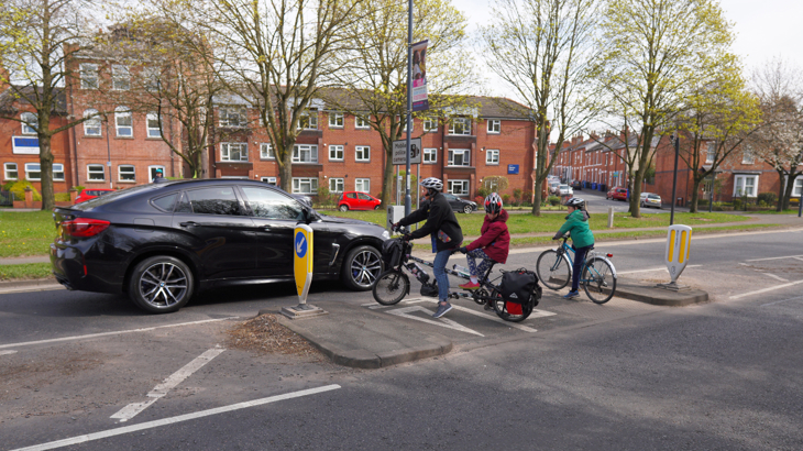 Kate riding her electric tandem with her child on the back. Her other child rides a bike beside them. The three are wearing helmets and waiting on the central reservation of a busy urban road.