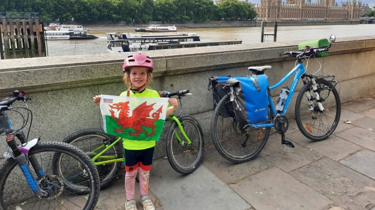 Emmer's four-year-old daughter proudly holds a Welsh flag beside the river Thames in London. The Houses of Parliament can be seen across the water and beside her are the family's luggage-loaded bikes.