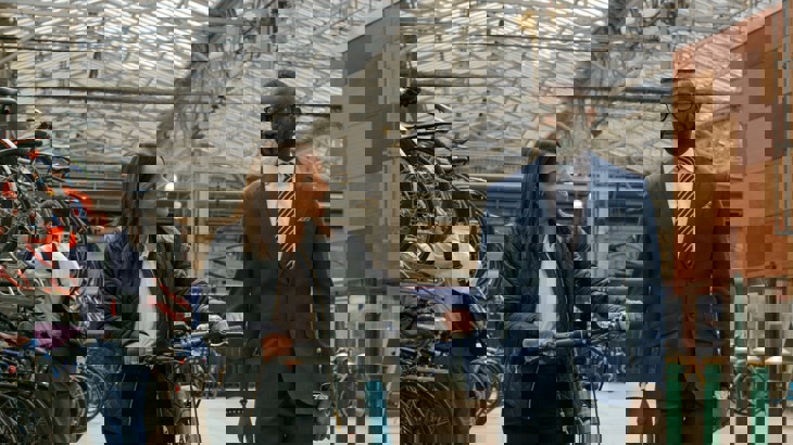 Two young professionals walking through a train station talking, one with a bicycle