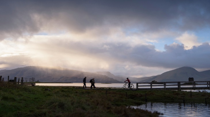 Two people walking and one cycling in silhouette against a dramatic cloudy backdrop on National Route 78 in Scotland