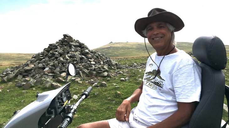 Shail, the author, on a mobility scooter on Dartmoor. Shail is smiling, wearing a Dartmoor t-shirt, shorts and hat. The day looks bright and warm.
