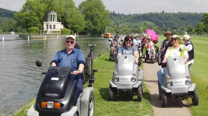 A large group of disabled ramblers making a journey along a gravel path and lawn beside the river Thames. All are laughing and smiling. It looks a warm, bright day. In the background are rolling green hills covered in trees. There is an island in the river with a small Palladian style building.