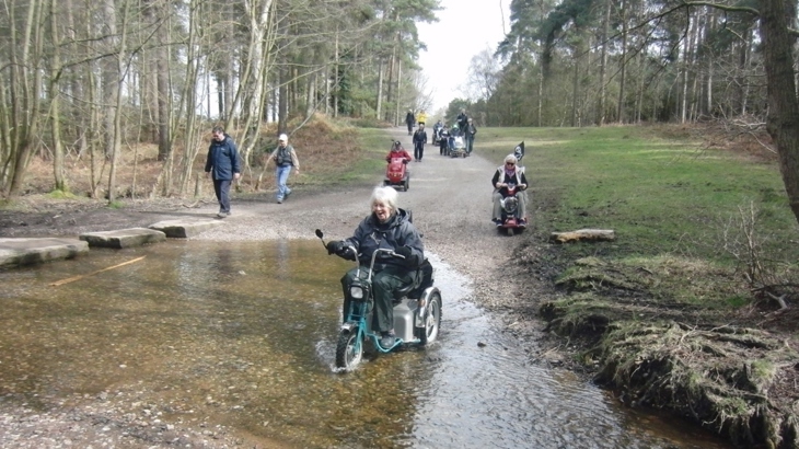 A disabled ramblers group on a wide stony path in woodland. In the foreground is a woman driving her scooter though a ford. She is laughing and smiling. All are in coats, it looks like a bright cold day.