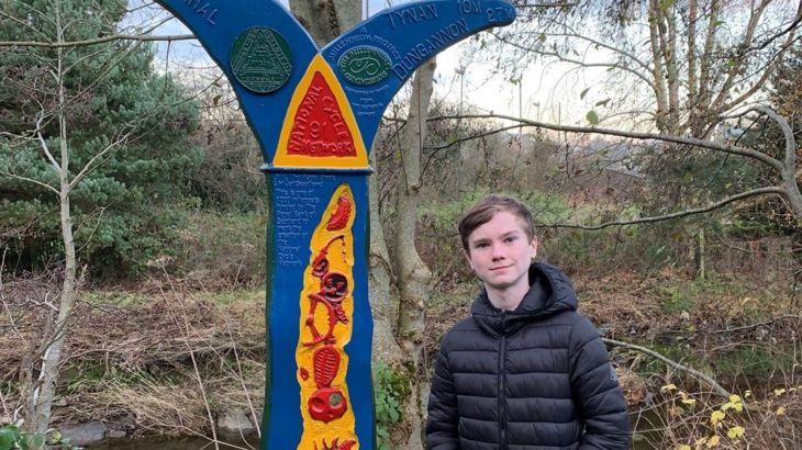 A teenage boy stands beside a brightly painted Millennium Milepost alongside a river.