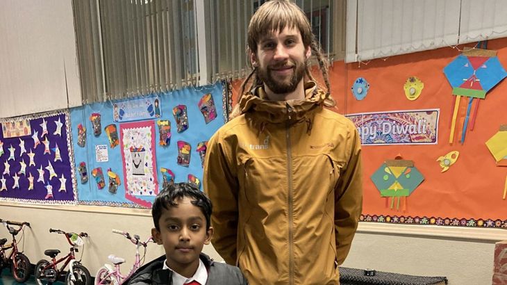 A young man wearing a  Walk Wheel Cycle Trust coat stands beside a young boy inside a school hall.
