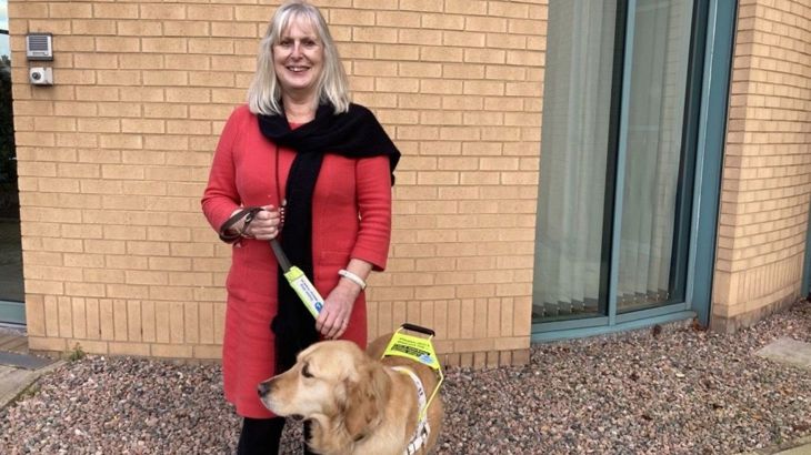 A woman wearing a red coat and black coat is standing outside holding the lead of her Golden Retriever guide-dog.