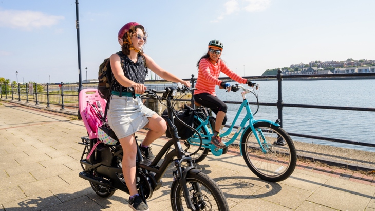 Two women riding e-bikes in sunny weather by a residential waterfront.