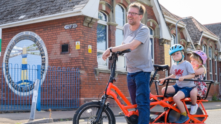 A man on a stationary e-bike in front of a school, with two children on the back, smiling, wearing helmets.