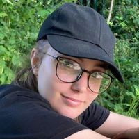 A closeup of a young person with glasses and a black baseball hat on smiling at the camera